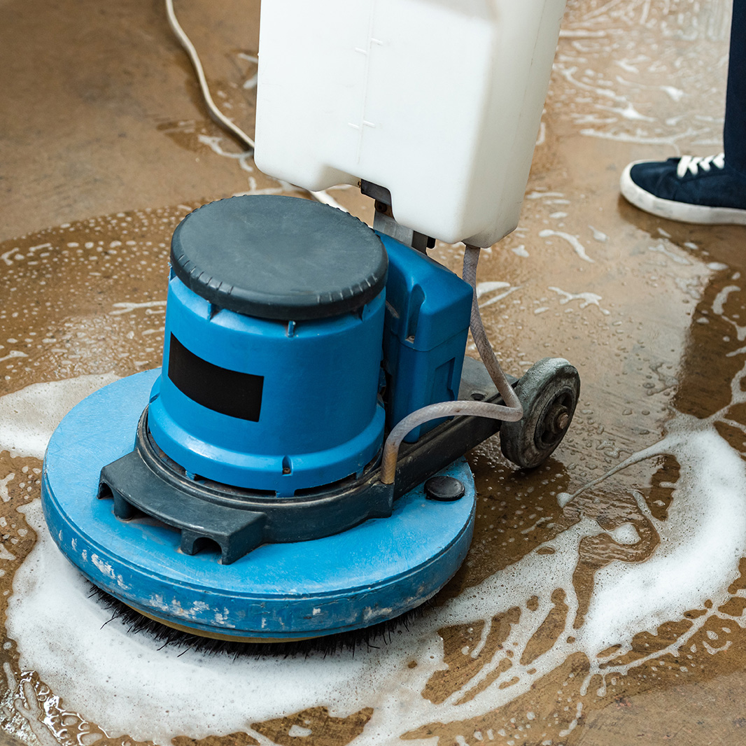 Eine Reinigungsmaschine schäumt den Baustellenboden ein, Beine einer Person in blauen Schuhen sichtbar, Wasser und Schaum auf dem Boden.