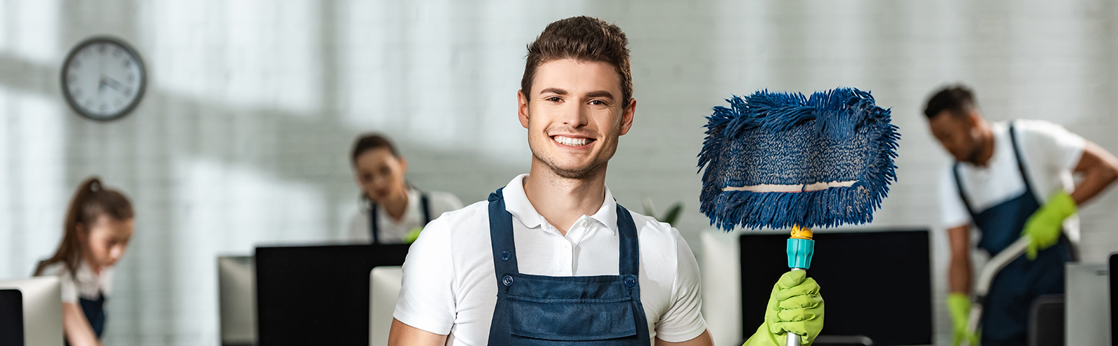 Ein lächelnder Reinigungskraft in blauem Overall mit grünen Handschuhen hält einen Staubwedel, Kollegen reinigen im Hintergrund.