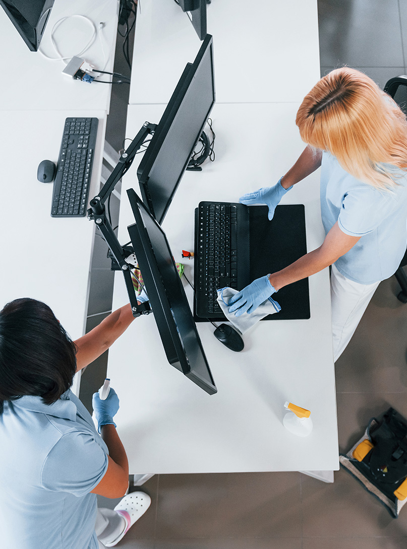 Zwei Reinigungskräfte in blauen Uniformen und Handschuhen reinigen einen Bürotisch mit mehreren Monitoren, einem Laptop und einer Tastatur, während Reinigungsmittel und ein Staubsauger sichtbar sind.