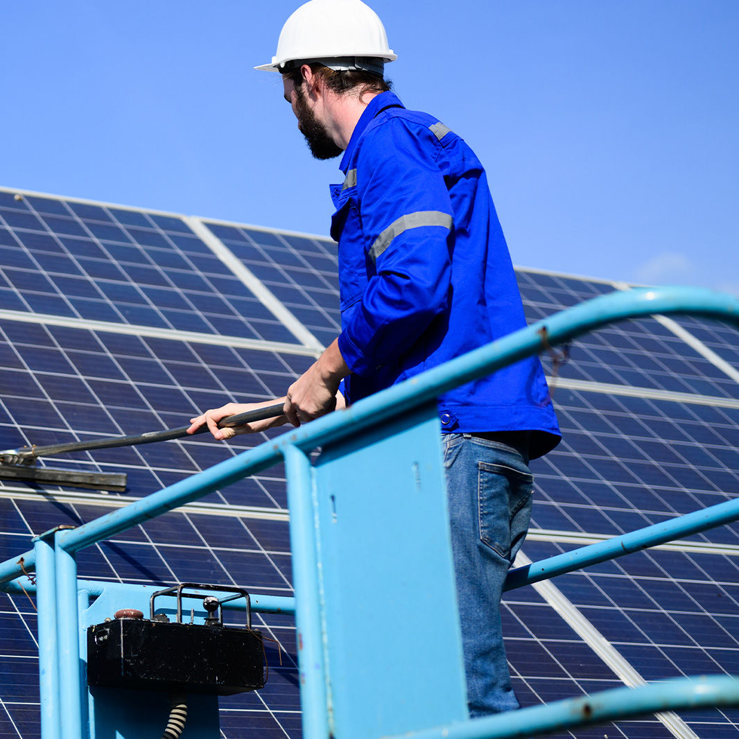 Ein Techniker in blauem Overall und Helm reinigt Solarmodule mit einem Schrubber von einer Plattform unter klarem Himmel.