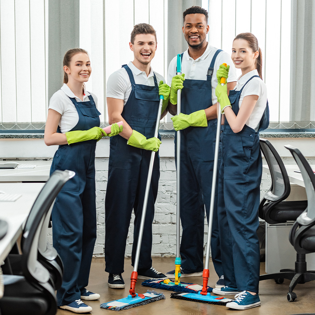 Vier Reinigungskräfte in blauen Overalls mit grünen Handschuhen stehen mit Mopps in einem Büro mit Schreibtischen und Stühlen.