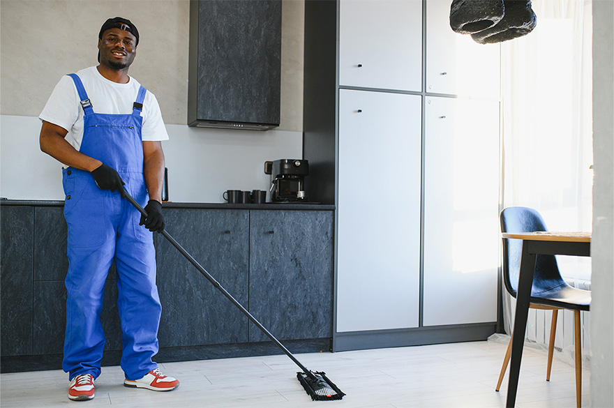Ein Reinigungskraft in blauer Arbeitskleidung und Handschuhen wischt mit einem Mopp den Boden einer modernen Küche mit schwarzen und weißen Schränken, einem Kaffeeautomaten und einem Tisch mit Stuhl neben einem Fenster.